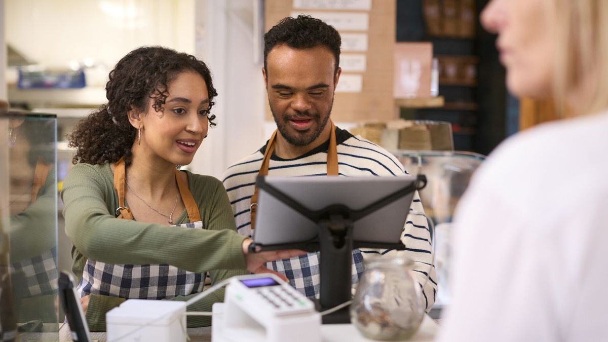 Woman Working In Food Shop Training Man With Down Syndrome How To Use Checkout